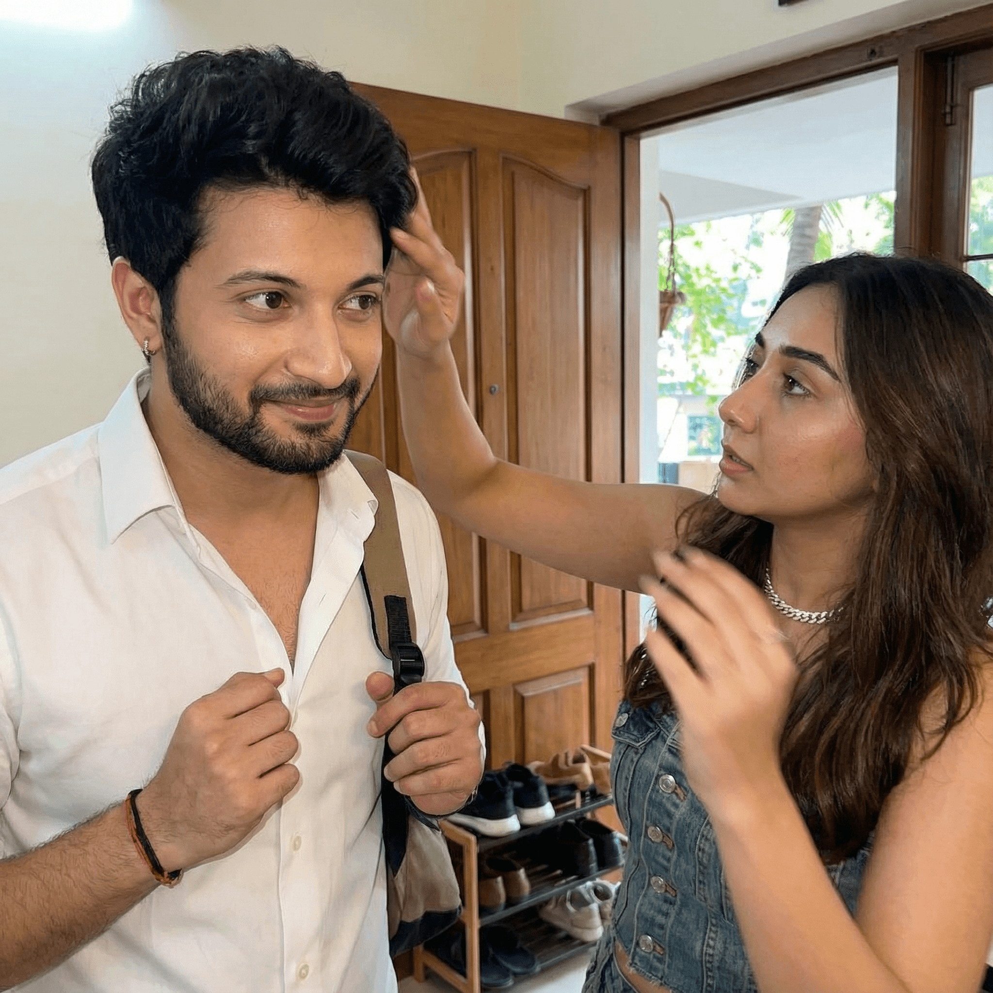 A close-up, fast-paced shot of an Indian couple at their front door. The man is looking slightly impatient to leave, while the girl is quickly adjusting his shirt collar or smoothing down his hair. Her expression is focused and caring, and his is appreciative, capturing the quick, close intimacy of commute prep.
