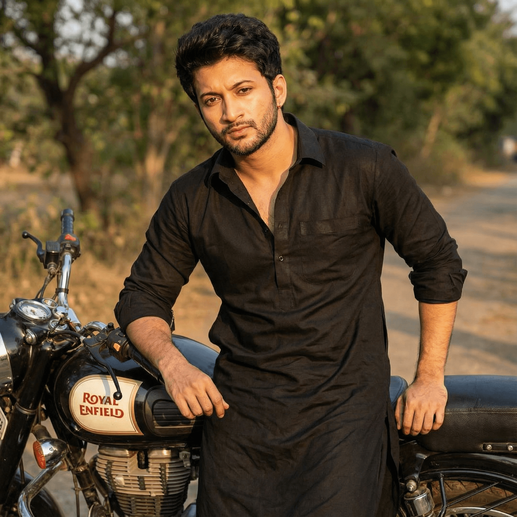 A confident portrait of a man wearing a black Pathani suit with rolled-up sleeves, leaning against a classic Royal Enfield Bullet motorcycle. He has a rugged stubble and is looking intently at the camera. Lighting: Late afternoon "golden hour" sun.