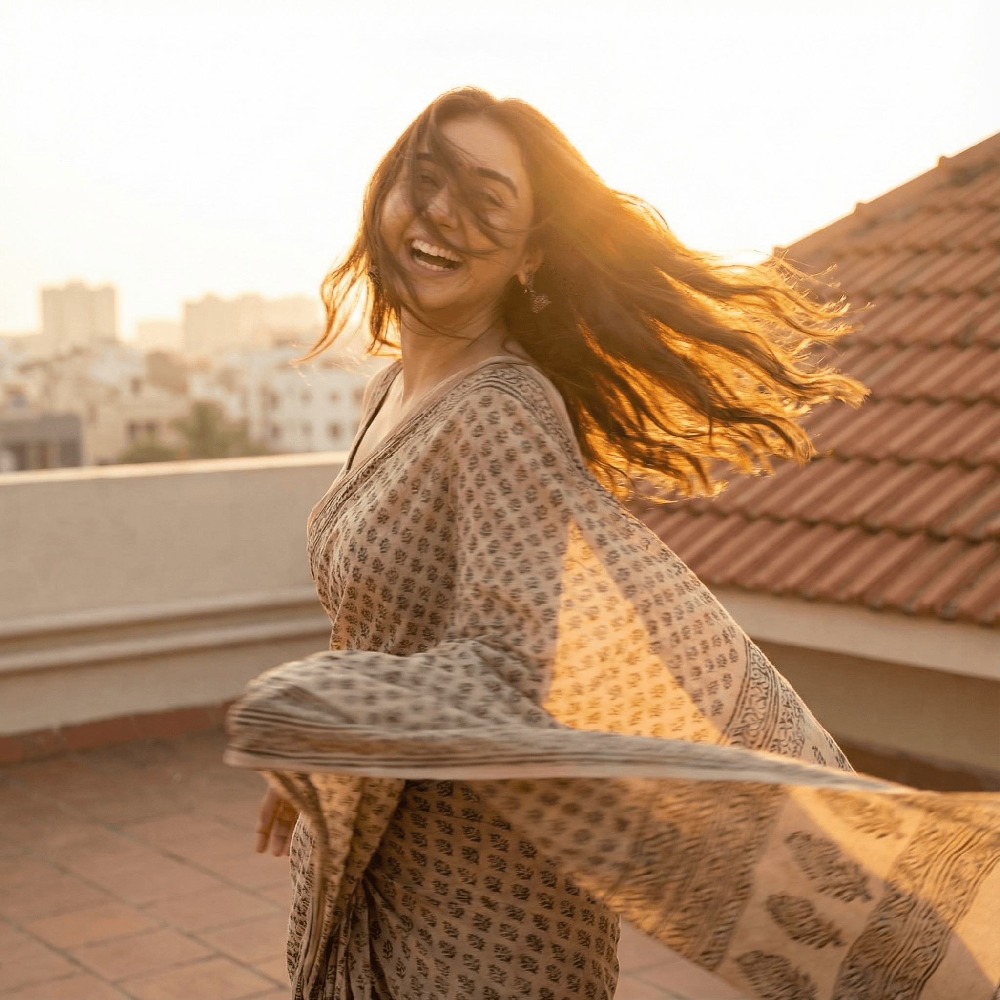 A candid, motion-blurred shot of a girl spinning around in a simple cotton saree on a rooftop terrace at sunset. She is looking back over her shoulder. Expression Cue: Her hair is flying across her face. She is laughing uncontrollably, looking breathless and free. Not a posed model smile. Lighting: Golden hour "halo" backlight.