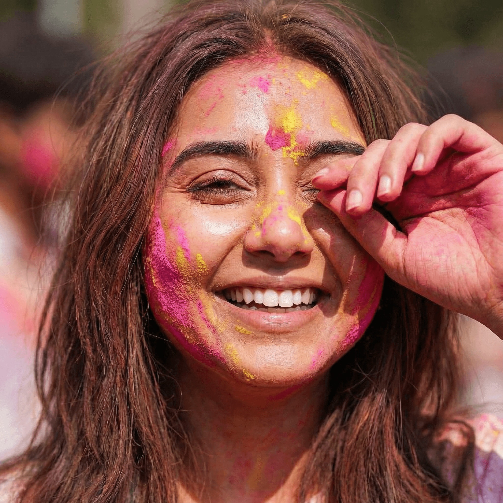 A close-up of a girl's face covered in pink and yellow Gulal. She is wiping color out of her eye with the back of her hand. Expression Cue: She is grinning broadly, showing white teeth against the color. One eye is squinting. She looks messy but radiant.