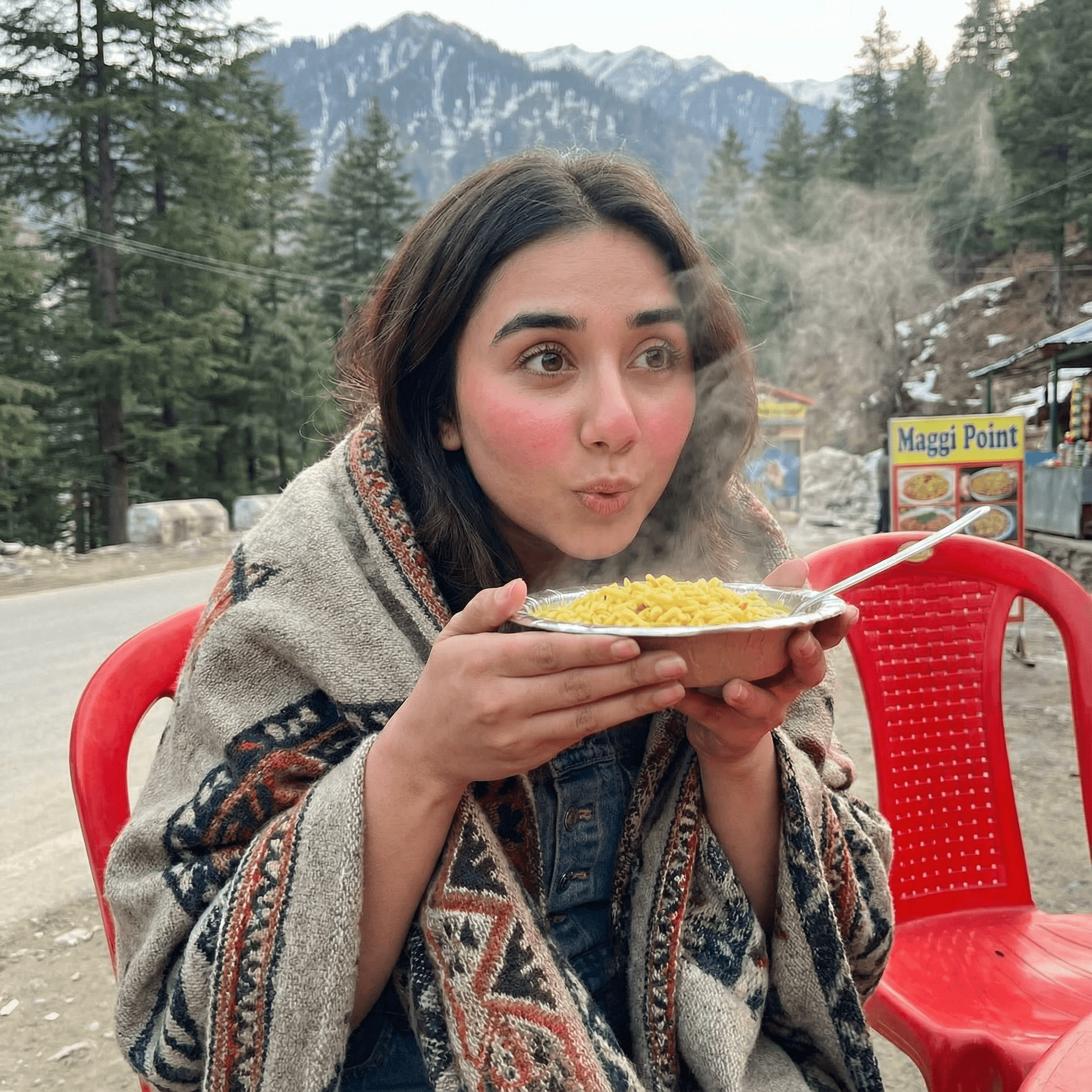 A cozy shot of a girl sitting on a plastic chair at a roadside mountain stall (Himachal), blowing on a steaming bowl of Maggi noodles. She is wrapped in a thick shawl. Expression Cue: Eyes focused on the food with "hungry anticipation." Cheeks flushed pink from the cold. A look of simple comfort.