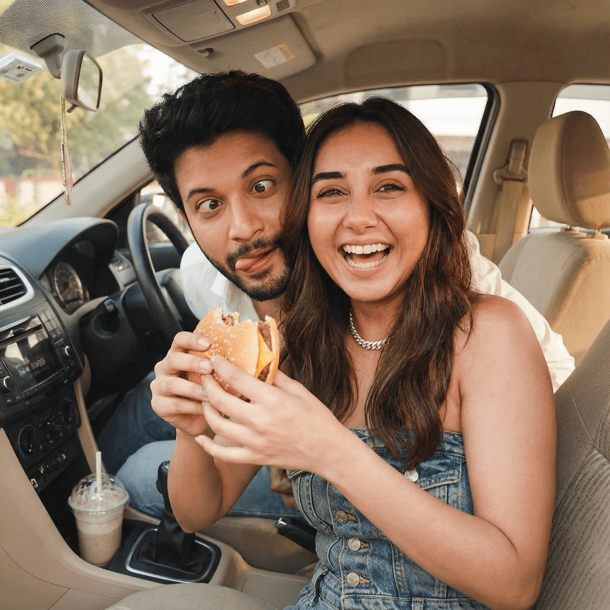 A playful, close-up shot of an Indian couple in their car. The girl is holding up a half-eaten burger or a large milkshake, smiling widely at the camera, while the man leans in from the driver's seat, making a goofy face. The interior of the car is cozy, conveying a fun, casual date.