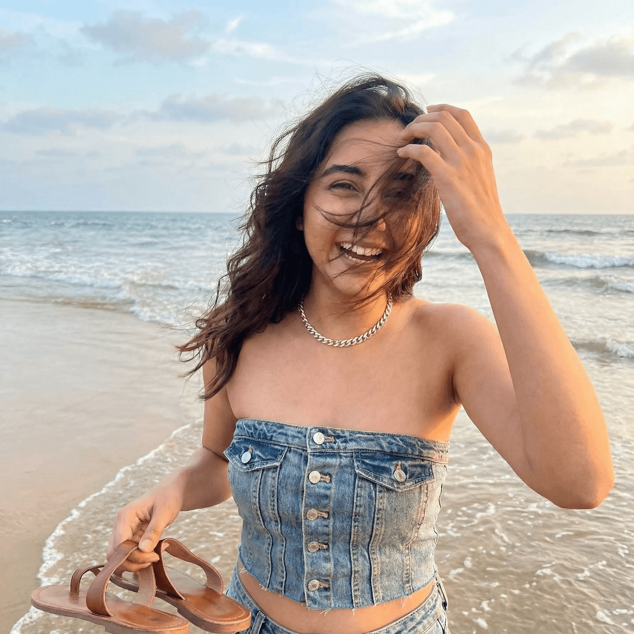 A girl standing on a beach with her feet in the water, holding her sandals in one hand. The wind is blowing her hair completely over her face. Expression Cue: She is brushing the hair away with a hand, laughing at the wind. Not a perfect hair flip—a messy, real, happy moment.